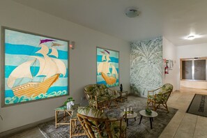 A room with a large sailboat painting, wicker furniture, a small table with bottles, and a patterned wall. - Apartamento Pet Friendly Beira-Mar no Guilhermina (Praia Grande)