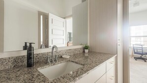 Un baño con encimera de granito, un lavabo, un espejo, una botella de jabón y una planta en maceta.
