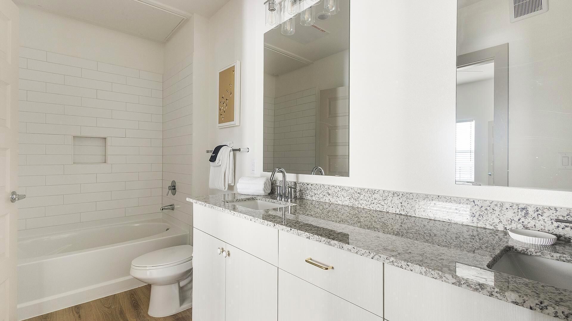 A bathroom with white subway tiles, a white toilet, a sink with a granite countertop, and a large mirror.