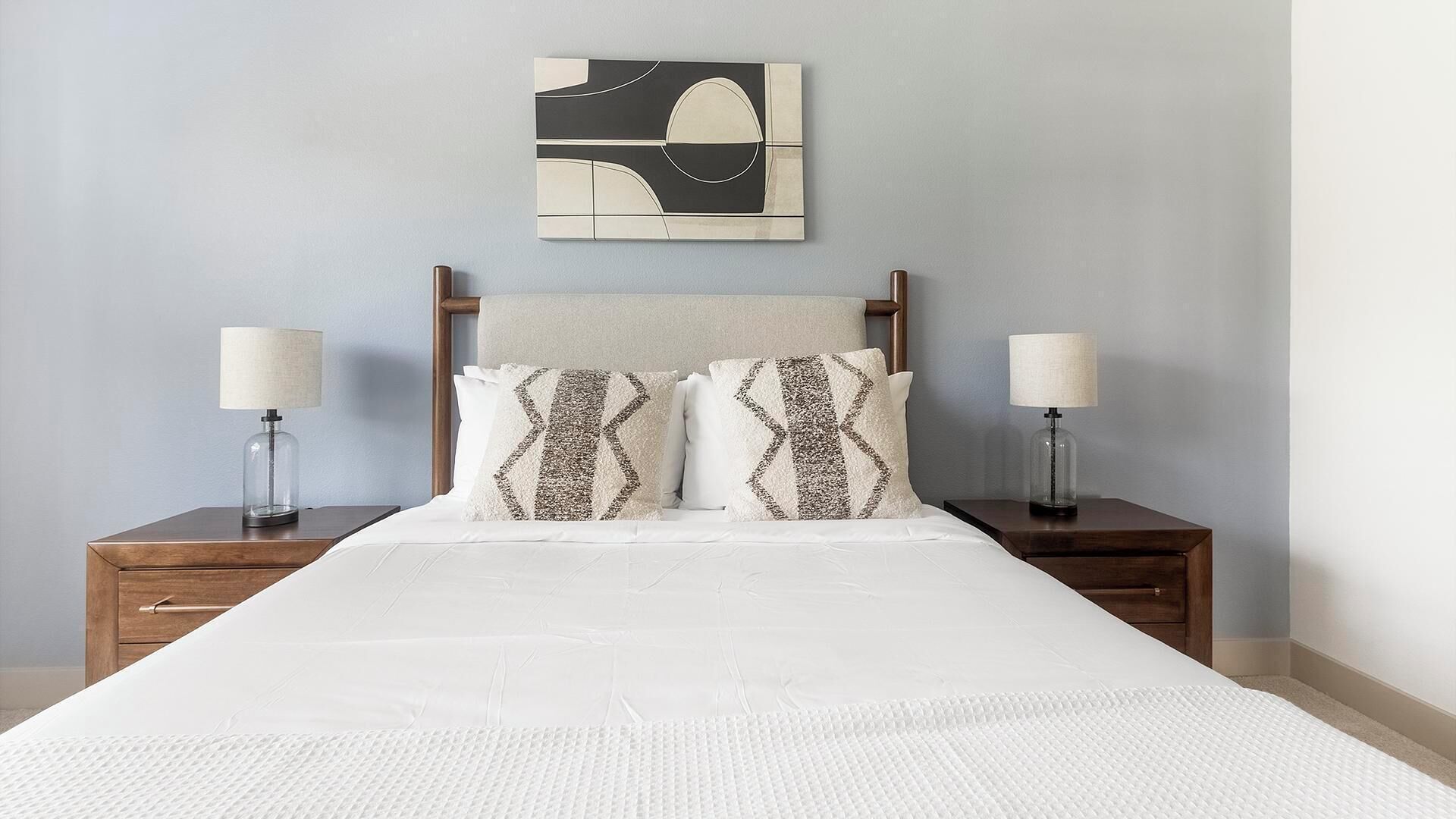 Un lit avec une couette blanche, deux coussins à motifs, une tête de lit et deux tables de chevet avec des lampes.