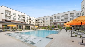 A swimming pool with lounge chairs and umbrellas, surrounded by multi-story buildings. - Landing | Modern 1BD, Courtyard, Yoga Studio (Houston)