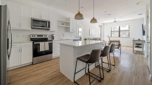 A modern kitchen with white cabinetry, a stainless steel refrigerator, a built-in oven, and a microwave. There are two pendant lights, a wooden island with bar stools, and a large window providing natural light. - Landing | Chic 2BD, Clubhouse, Gym (Austin)