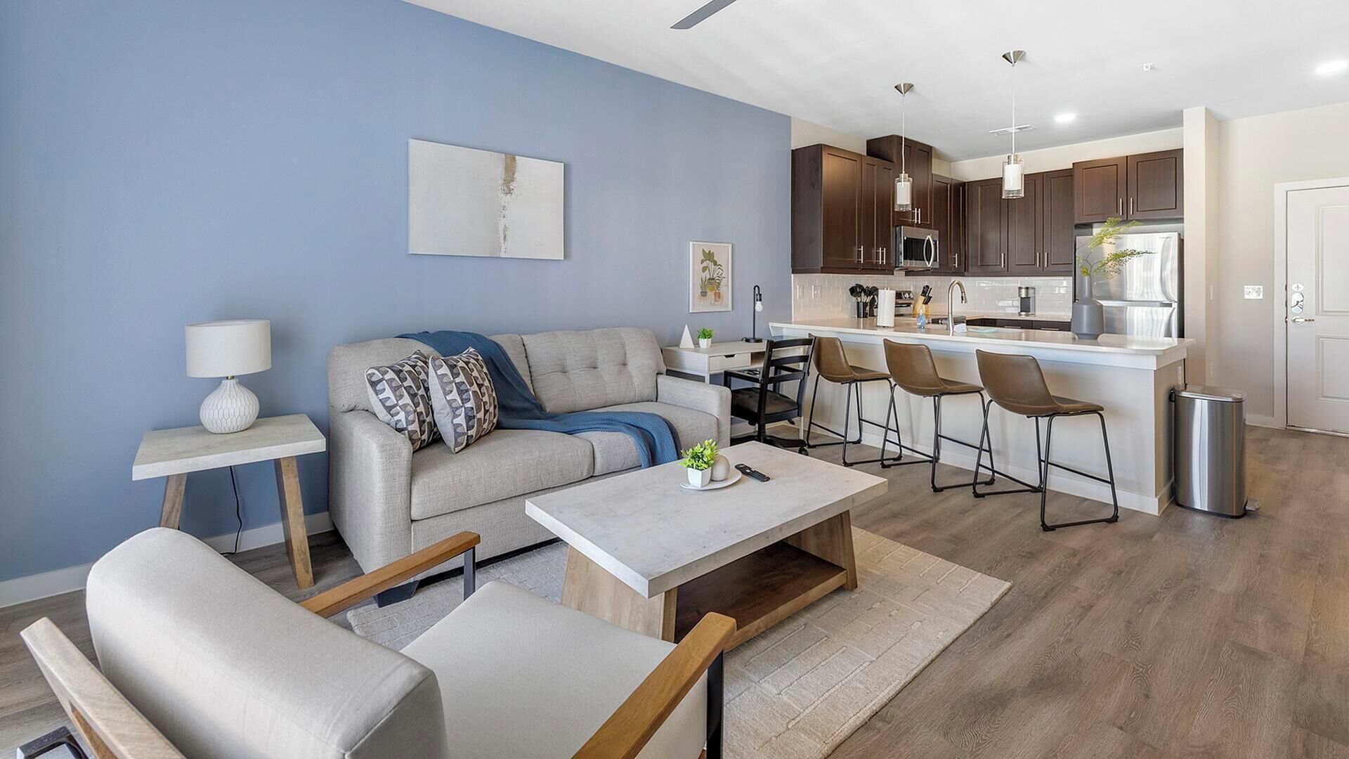Un salon moderne avec un canapé, une table basse et des tabourets de bar, une cuisine avec des meubles sombres et un plan de travail blanc, et un mur d’accent bleu.