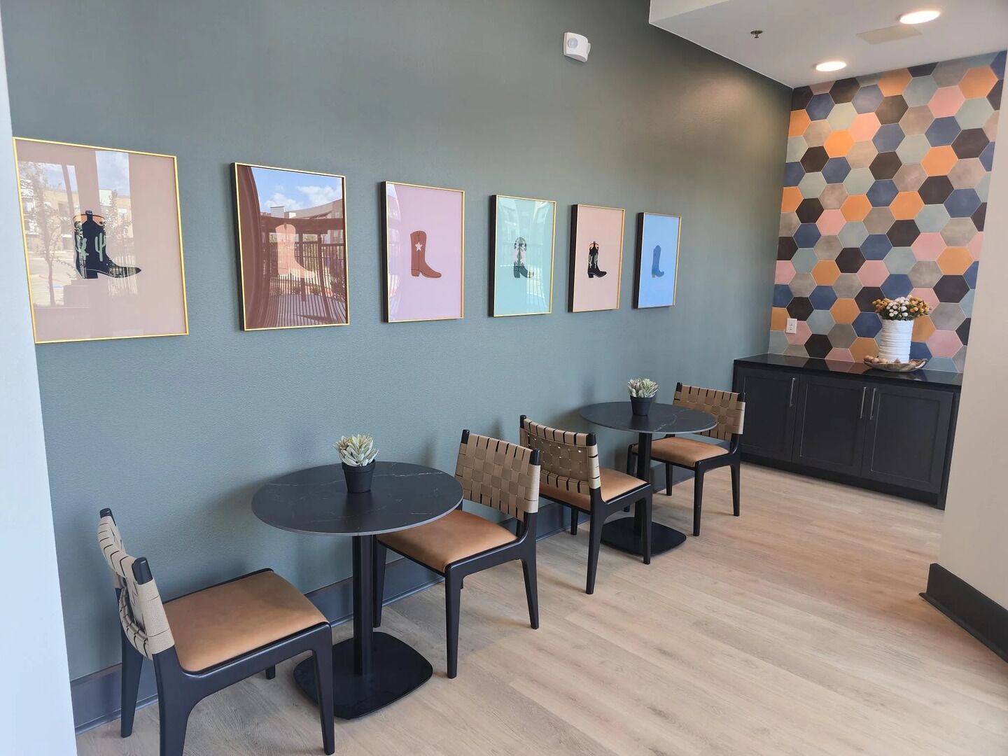 Un intérieur moderne avec un espace salon comprenant des tables rondes et des chaises rembourrées, un mur recouvert d’un papier peint à motifs hexagonaux et des œuvres d’art encadrées.