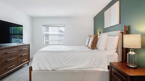 A hotel room with a bed, a dresser, a TV, a lamp, and a window with a view of buildings. - Landing | Modern 2BD, Gym, Clubhouse (Kissimmee)