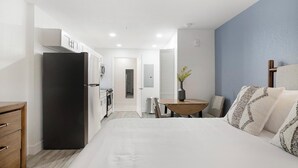 A hotel room with a bed, a desk, a chair, a small table with a vase, and a kitchenette. - Landing | Stylish Studio, Gym, Pool (Kissimmee)