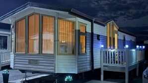 A mobile home with large windows, a porch, and a vehicle parked nearby. - Happy Days Caravan Park - Holiday Accommodation 21912 (Abergele)