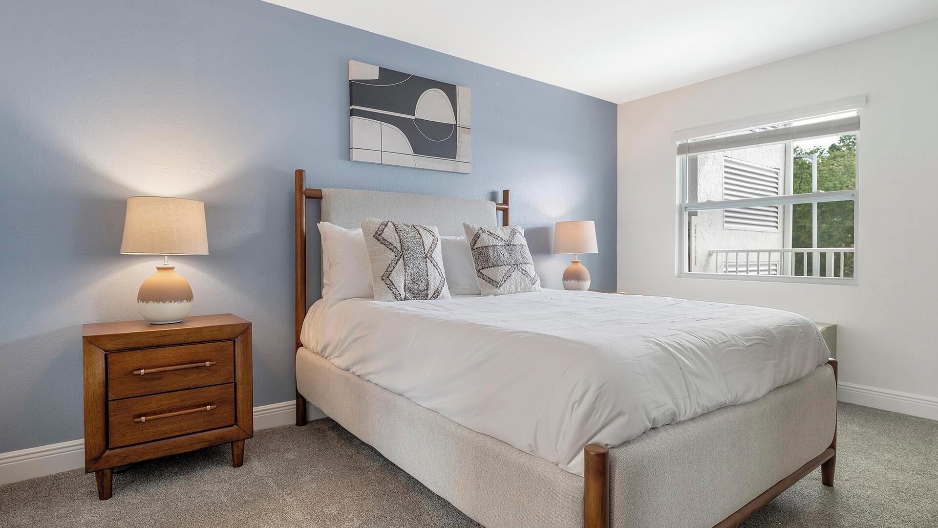 A bedroom with a bed, two bedside tables, a window, and a wall-mounted artwork.