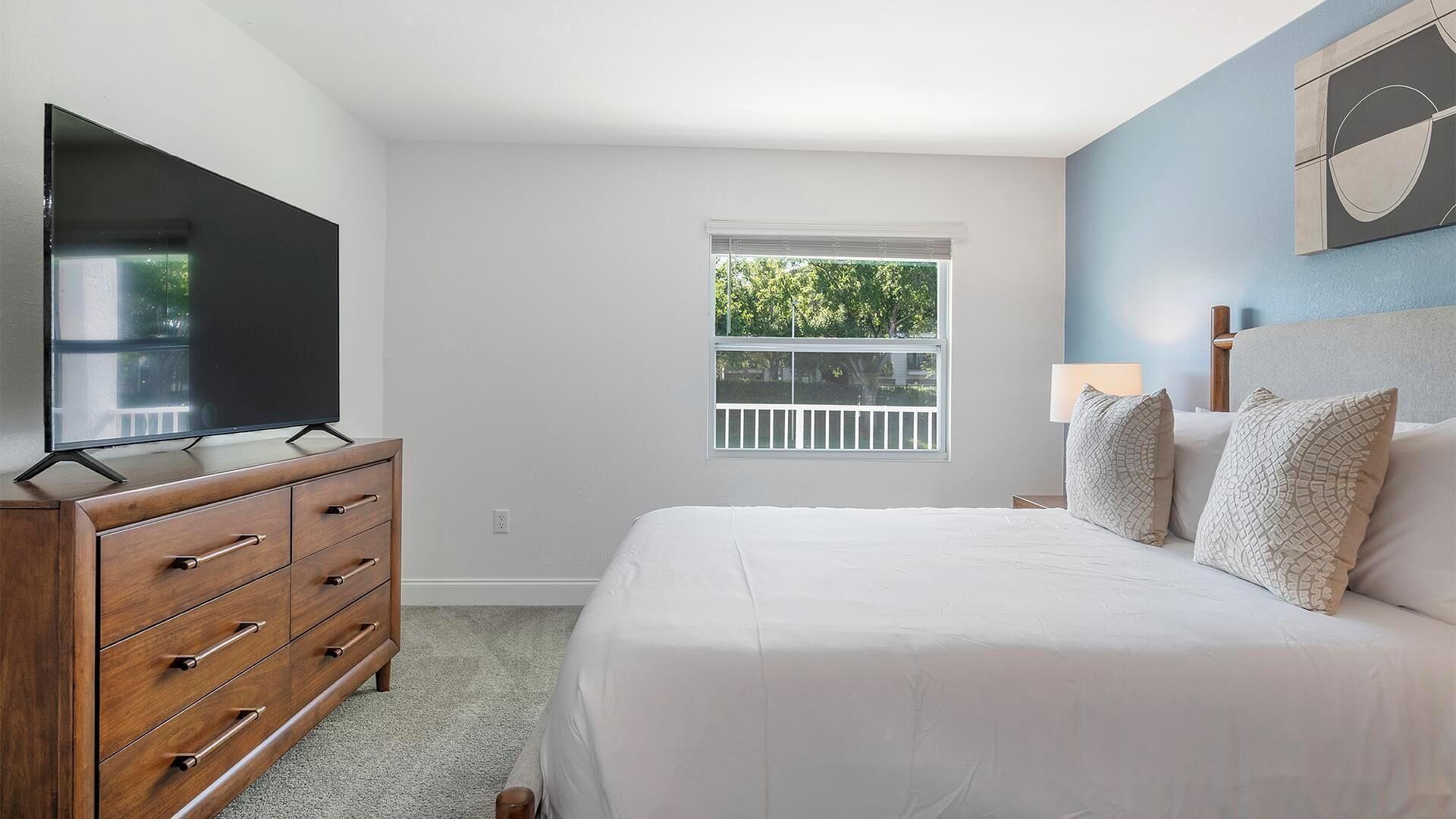 A bedroom with a bed, dresser, TV, window, and a view of greenery outside.