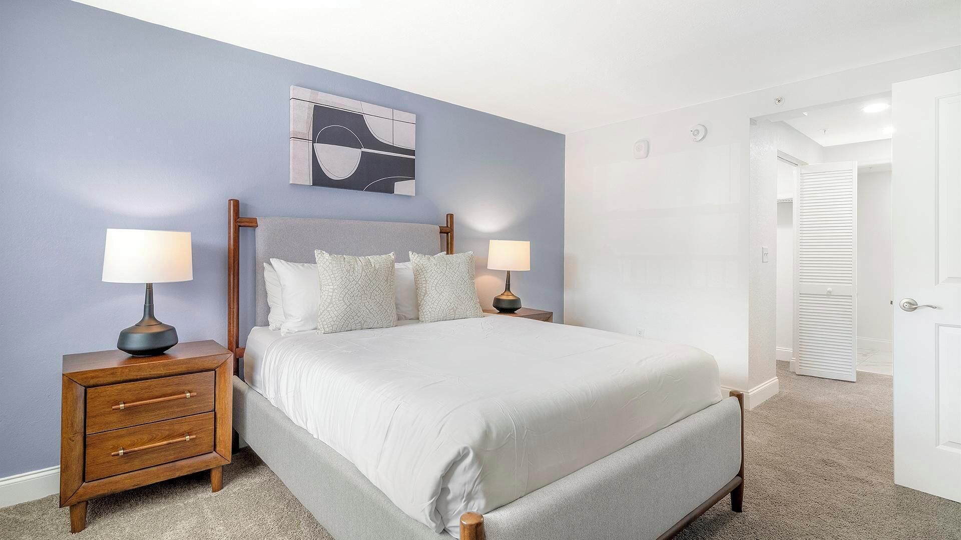 Um quarto com uma cama, duas mesas de cabeceira, um criado-mudo, uma obra de arte na parede e uma porta para um armário.