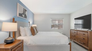 A bedroom with a bed, bedside tables, a dresser, a TV, and a window with blinds. - Landing | Stylish 2BD, Pool, Gym (Kissimmee)