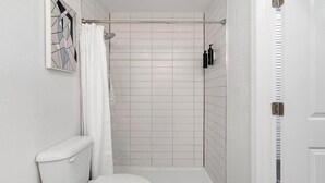 Un baño pequeño y blanco con ducha, inodoro y un estante en la pared con artículos de aseo.