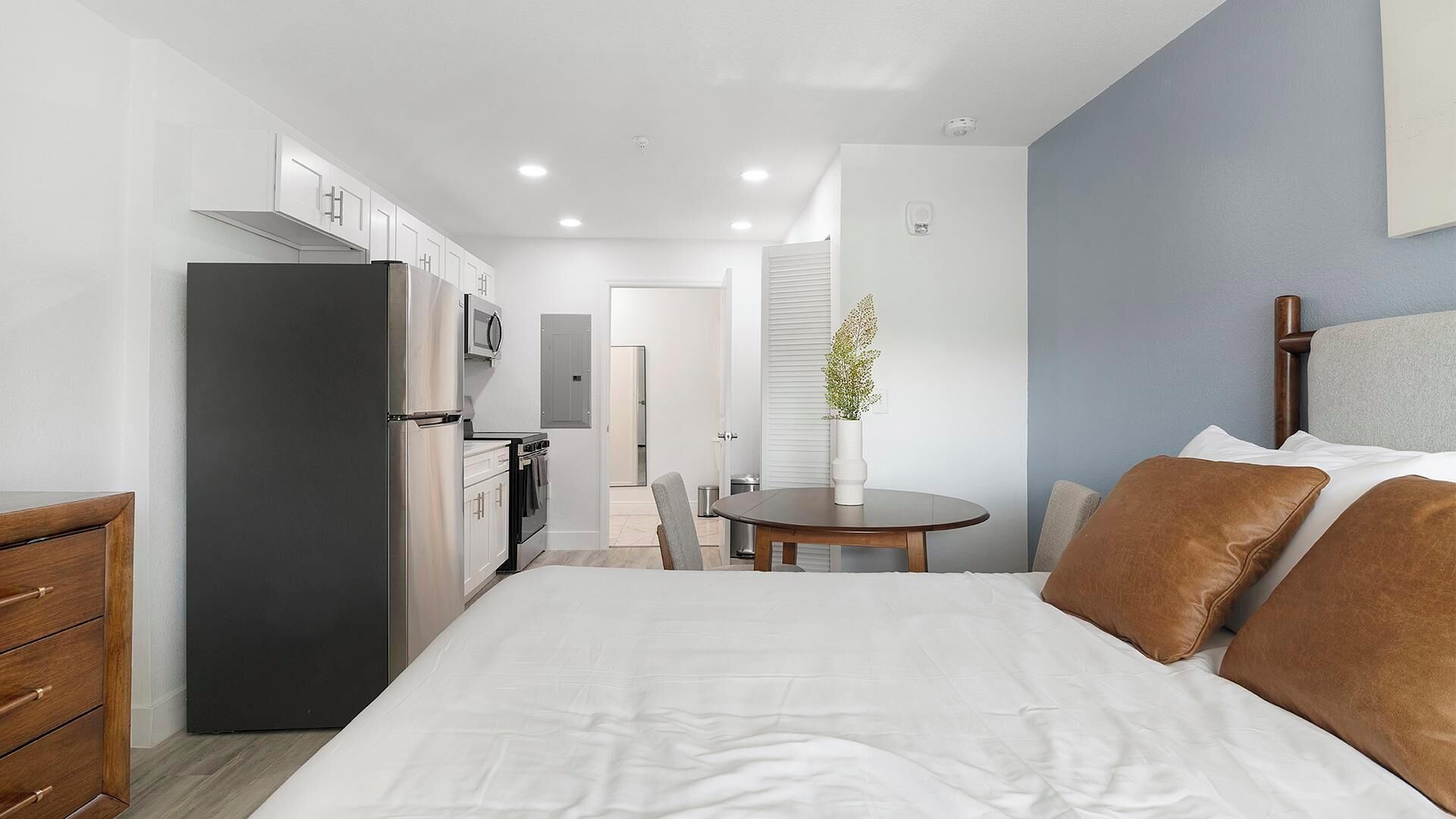 Ein Hotelzimmer mit einem Bett, einem Nachttisch aus Holz, einem Schreibtisch mit Stuhl, einer Küchenzeile und einem sichtbaren Badezimmer durch eine offene Tür.