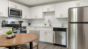 A modern kitchen with white cabinets, stainless steel appliances, a dining table with a plant, and a chair. - Landing | Beautiful 1BD, Pool, Clubhouse (Kissimmee)