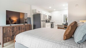 A bedroom with a bed, a TV, a wooden dresser, and a kitchenette in the background. - Landing | Stunning Studio, Pool, Clubhouse (Kissimmee)