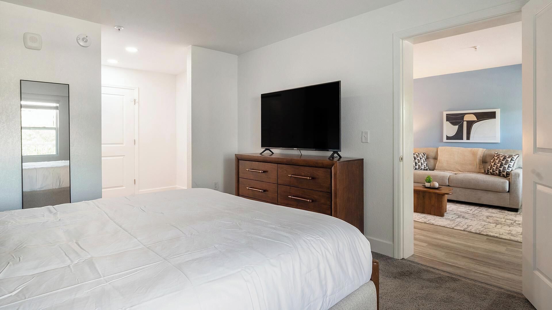 A bedroom with a bed, a dresser, a TV, and a door leading to a living area with a sofa and a coffee table.