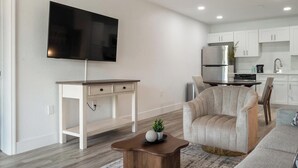 Een moderne woonkamer met een flatscreen televisie, een houten bureau, een comfortabele fauteuil en een keuken in de achtergrond.