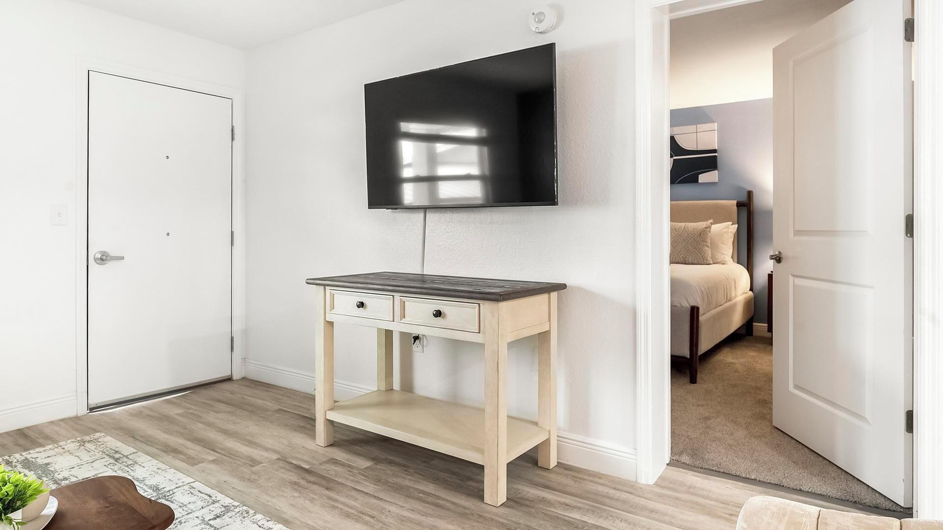 A room with a wooden desk, a mounted TV, and a bed in the adjacent room.