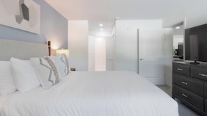 Ein Schlafzimmer mit Bett, Nachttischlampe, Kommode, Fernseher und einer Tür zu einem weiteren Raum.