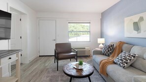Ein Wohnzimmer mit einer Couch, einem Sessel, einem Couchtisch, einem Fernseher und einem Fenster mit Blick auf Bäume.