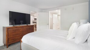 A bedroom with a bed, a wooden dresser, a flat-screen TV, and a door leading to a bathroom. - Landing | Beautiful 1BD, Gym, Pool (Kissimmee)