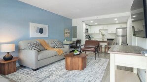 A living room with a sofa, coffee table, and a TV. There is a kitchen visible in the background. - Landing | Amazing 1BD, Pool, Gym (Kissimmee)