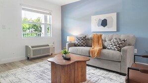 A living room with a grey sofa, a wooden coffee table, a radiator, and a window with blinds. - Landing | Amazing 1BD, Pool, Gym (Kissimmee)