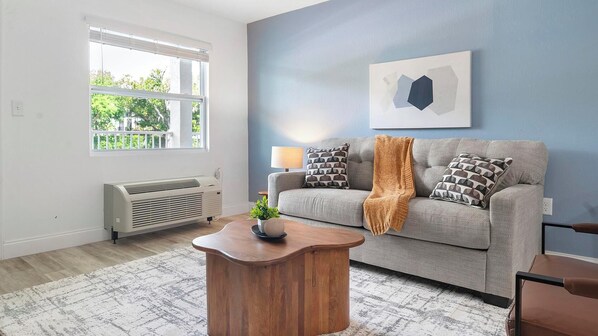 A living room with a grey sofa, a wooden coffee table, a radiator, and a window with blinds. - Landing | Beautiful 1BD, Gym, Pool (Kissimmee)