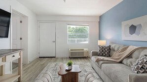 A living room with a sofa, a coffee table, a TV, a door, a window, and an air conditioner. - Landing | Incredible 1BD, Pool, Gym (Kissimmee)
