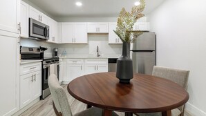 A modern kitchen with white cabinetry, a stainless steel refrigerator, a microwave, and a stove. There's a round wooden table with chairs and a vase with flowers. - Landing | Incredible 1BD, Pool, Gym (Kissimmee)