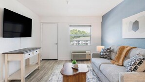 A modern living room with a flat-screen TV, a sofa, a coffee table, and a window with a view of greenery. - Landing | Stunning 1BD, Clubhouse, Pool (Kissimmee)