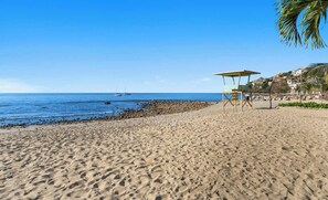 Una playa con un puesto de salvavidas, costa rocosa y un cielo azul despejado.