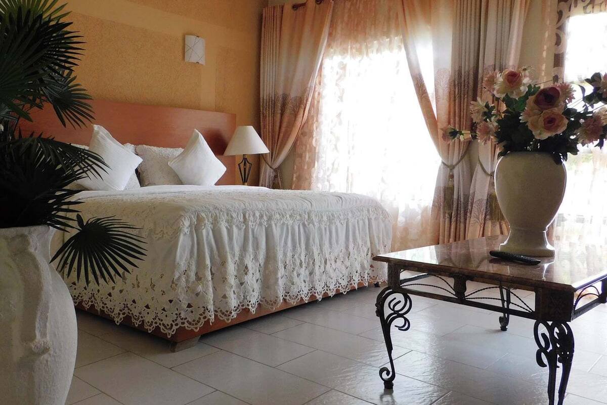 A hotel room with a bed, a bedside table, a lamp, a large vase with flowers, and a potted plant.