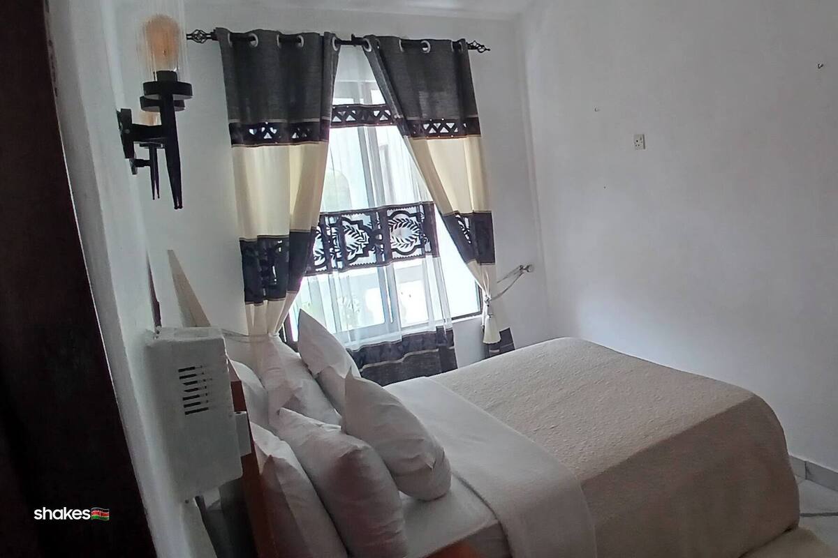 A small, simple room with a bed, a window with curtains, a wall-mounted air conditioning unit, and a power outlet.
