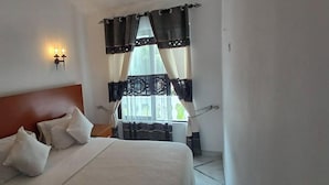 A bedroom with a bed, pillows, a headboard, a window with curtains, and wall-mounted lights. - Kigotho Hotel & Apartments (Mombasa)