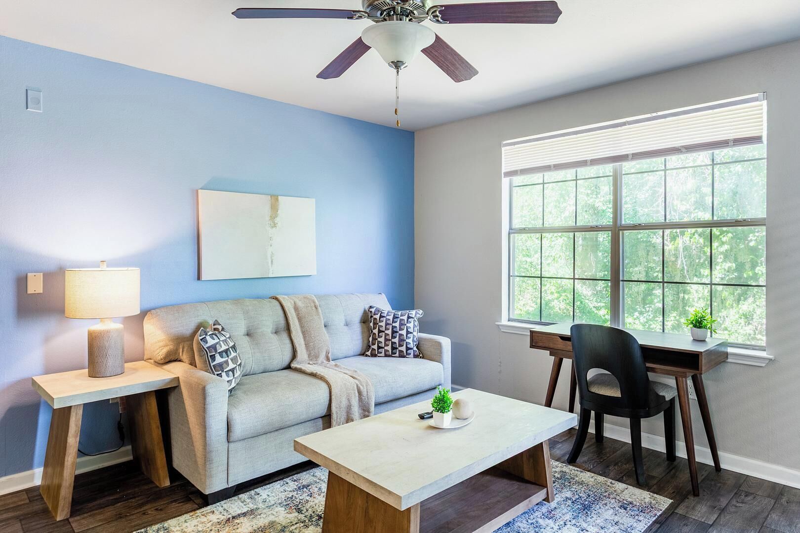 A living room with a sofa, coffee table, and a desk with a chair. There is a ceiling fan, a window with blinds, and a plant on the desk.