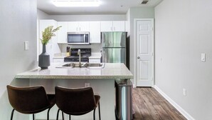 Una cocina compacta con armarios blancos, microondas, estufa, refrigerador y pileta. Hay dos sillas y un bote de basura.