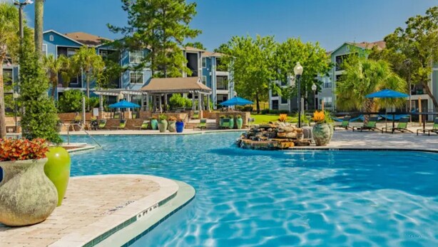 A swimming pool with a water feature, surrounded by lounge chairs and umbrellas, with buildings in the background. - Landing | Spectacular 2BD, Game Room, Clubhouse (Tallahassee)