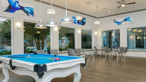 Uma mesa de bilhar com bolas, um ventilador de teto e luminárias modernas numa sala com grandes janelas e vista para o exterior.