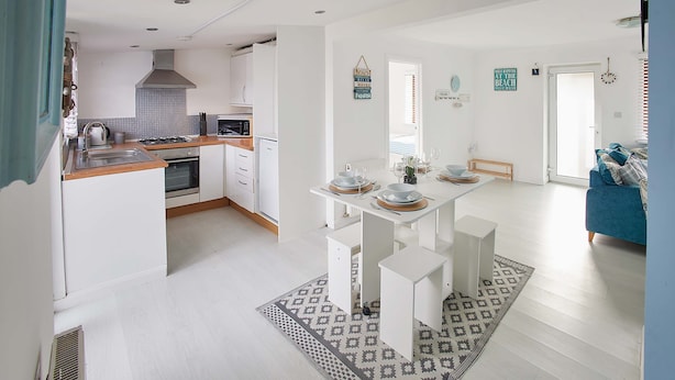 Eine moderne Küche mit weißen Schränken, ein Essbereich mit einem weißen Tisch und Bänken sowie ein Wohnzimmer mit einem blauen Sofa.