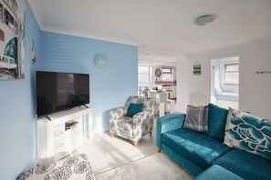 Un salon avec un téléviseur, un fauteuil à motifs, un canapé bleu et une cuisine en arrière-plan.