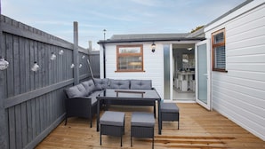 Eine Terrasse mit einer Couch, einem Tisch und Stühlen, ein Holzterrasse, ein grauer Zaun und ein weißes Gebäude mit Fenster und Tür.