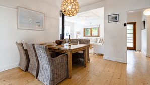 Uma área de refeições com uma mesa de madeira, cadeiras de vime, um lustre e um sofá branco ao fundo.