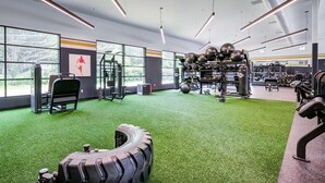 A gym with exercise equipment, a large tire, and a view of trees through the windows. - Landing | Chic 1BD, Yoga Studio, Gym (New Haven)