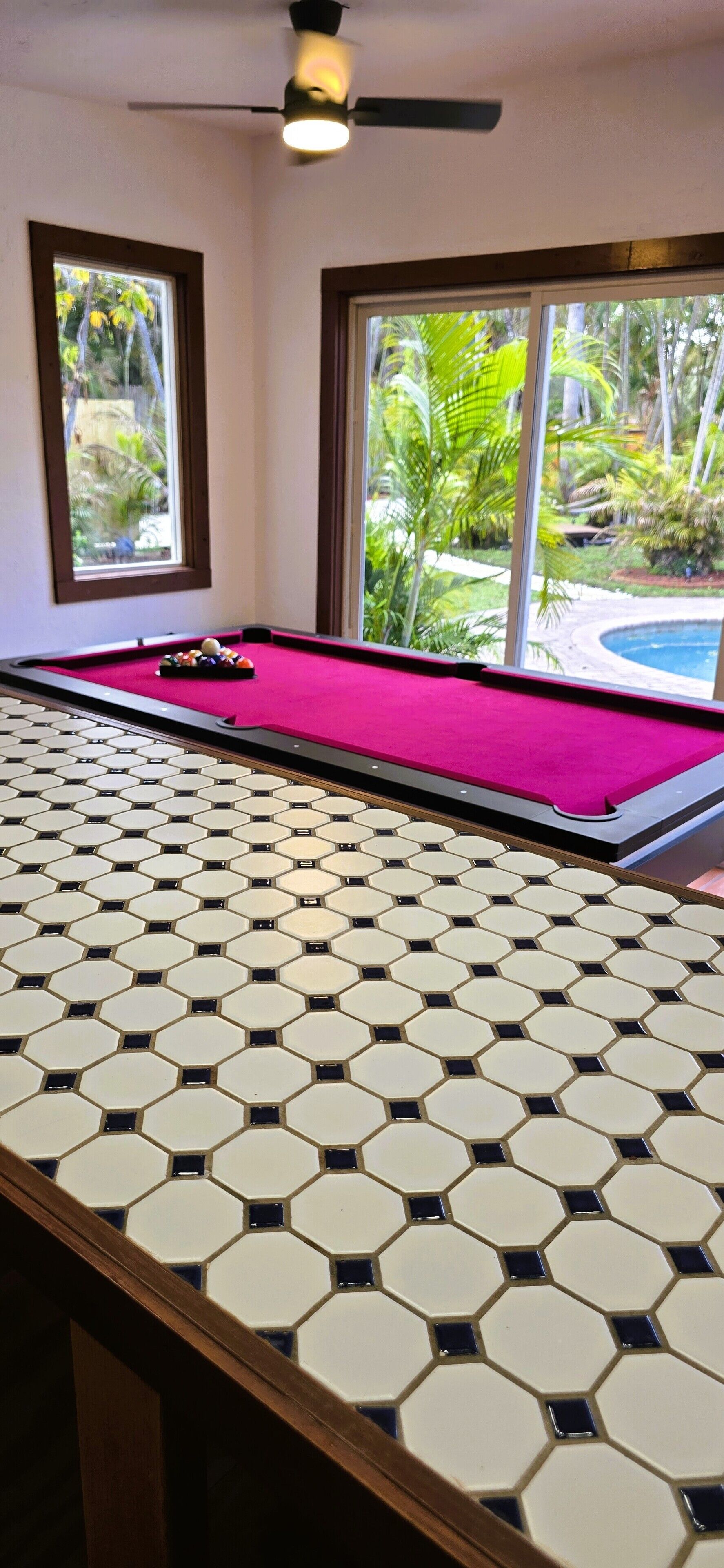 Une table de billard ornée d’un motif hexagonal en carreaux, recouverte d’un drap de feutre rouge, et offrant une vue sur une piscine et la verdure environnante grâce à de grandes fenêtres.