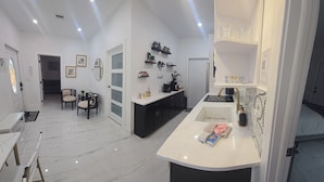 Een moderne keuken met een wit aanrecht, zwarte kastjes en een ingebouwde oven. Er is een eethoek met een tafel en stoelen, en een gang met een deur en een spiegel.