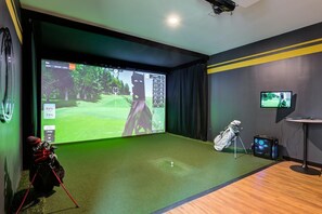 A room with a large screen displaying a golf game, a golf bag, and a tripod with a golf club.