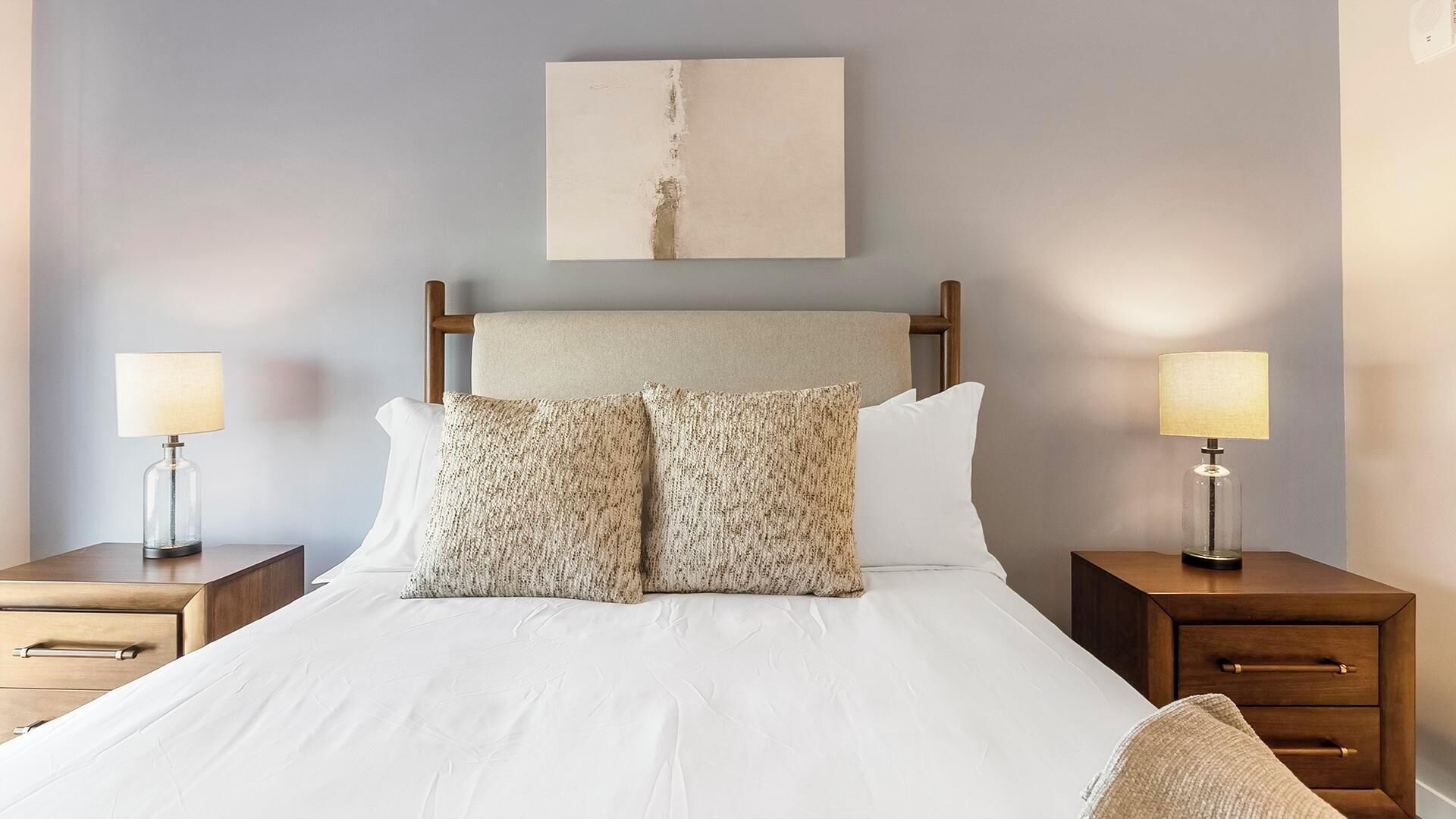Cama bem arrumada com duas almofadas, cabeceira de madeira e duas mesas de cabeceira com abajures.