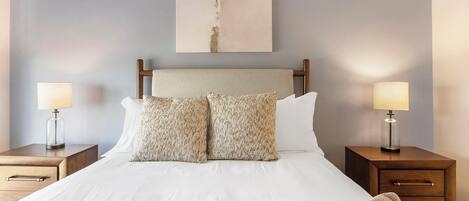 Cama bem arrumada com duas almofadas, cabeceira de madeira e duas mesas de cabeceira com abajures.