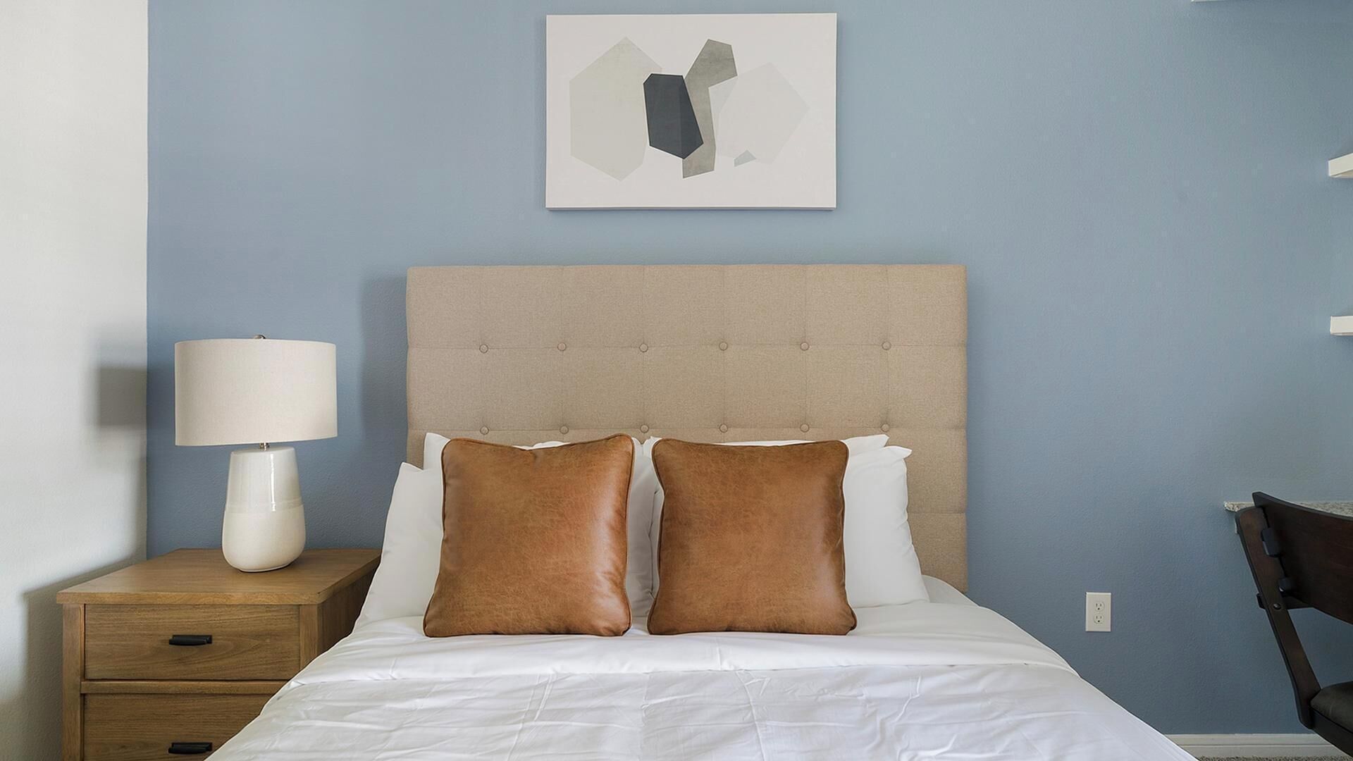 A bedroom with a tufted headboard, two pillows, a bedside table with a lamp, and a wall-mounted artwork.